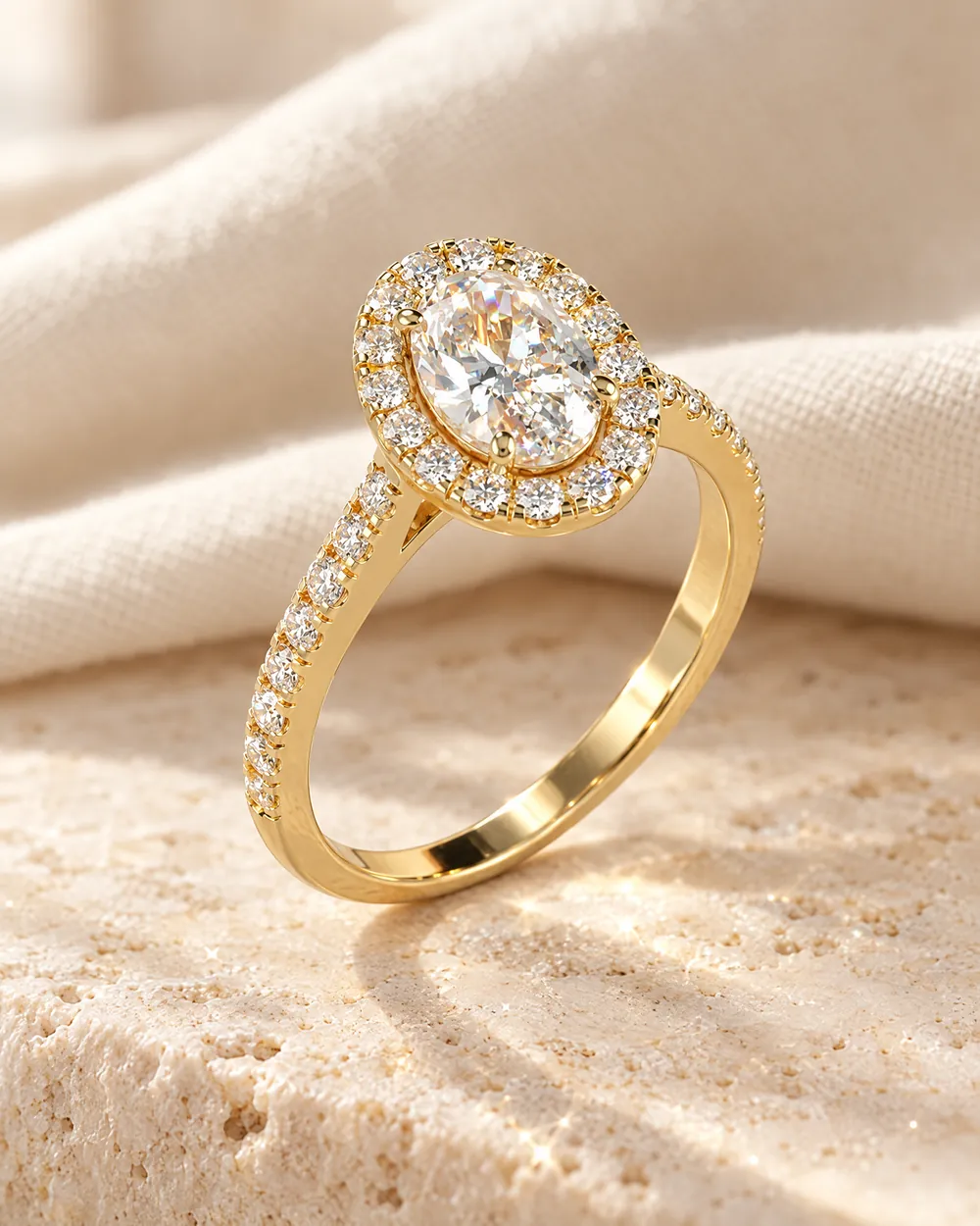 Verlobungsring Kelly: Diamant im Ovalschliff mit Halo und Pavé-Schiene, Gelbgold, auf hellem Travertin
