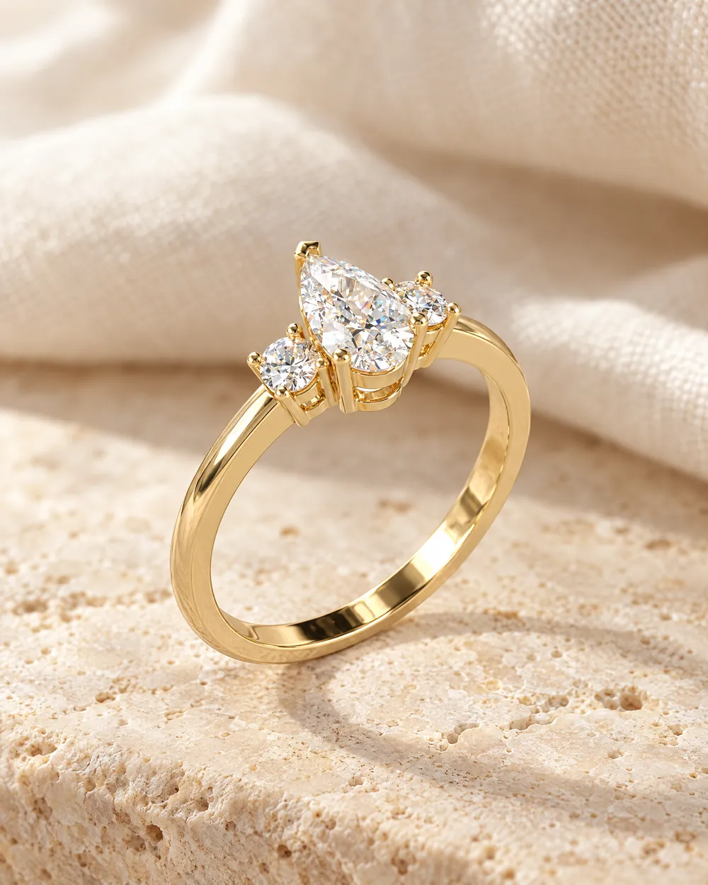 Verlobungsring Trinity: Diamant im Tropfenschliff mit zwei runden Diamanten, Gelbgold, auf hellem Travertin